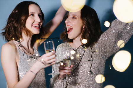 Portrait of twin sisters in shiny evening dresses celebrate with glasses of sparkling wine. they...