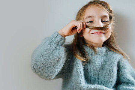 Smiling satisfied little girl in a blue knitted sweater playing with her hair, making fake mustache.