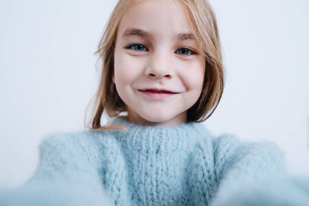 Portrait of a cheerful little girl in a blue knitted sweater over white. She has extended her hands in a selfie fashion. Close up.の写真素材