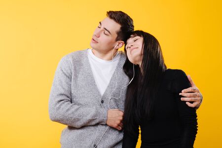 Young happy couple leaning on each other, listening to the music over yellow background. They are immersed in a feeling, their eyes are closed. His arm around her shoulder.の写真素材