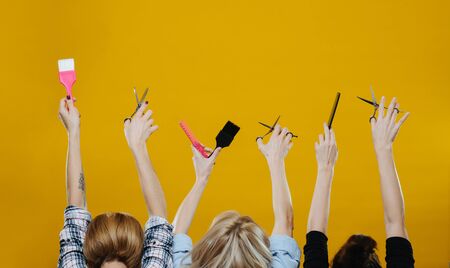 Women with their hands in the air holding hairdresser tools, such as brushes, scissors and combs over yellow backgound.の写真素材