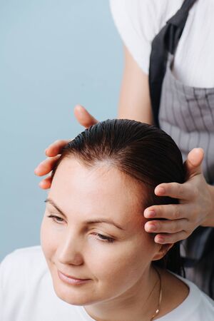 Focused blonde female hairdresser is preparing client's wet sleek brown hair for trimming. Over blue background. Close up. Cropped, customer's head only. Customer is happy, peaceful, and relaxed.の写真素材