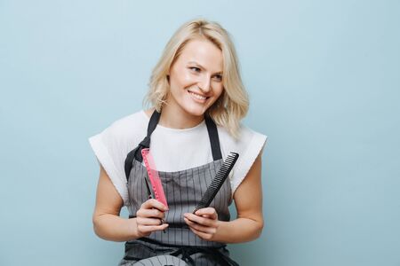 Portrait of a stylish blonde female hairdresser in apron with combs and scissors in hands over blue background.の写真素材