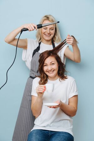 Blonde female hairdresser in apron using curling iron on client's brown hair, while customer drinking tea.の写真素材