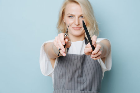Playful smirking stylish blonde female hairdresser in apron pointing at you with comb and scissors...