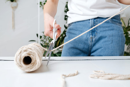 Woman measuring piece of the rope from the coil for macrame weaving with big tailor scissors. She's...