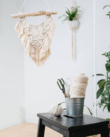 Composition of cotton rope coil in a bucket on stool along with two pairs of scissors. Macrame tapestry and pot in a holder hanging from a ceiling.の写真素材