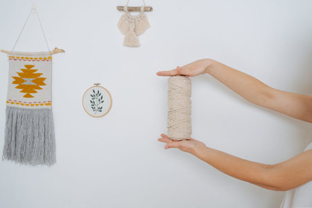 Woman hands holding rope coil next to macrame tapestries and little piece of handwork hanging on the...