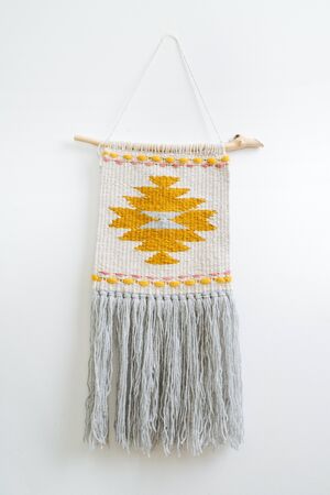 Macrame tapestry with yellow-colored patten hanging on the white wall of a room. Pattern contains yellow and pink dots and angular flower-like shape in the middle.の写真素材