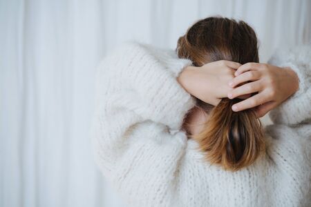 Brunette little girl in white fluffy knitted sweater putting her hair in a pony tail. At home, in front of a curtain. From behind.の写真素材