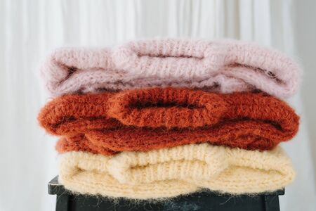 Three folded multi-colored knitted sweaters lie on a stool in front of a white curtain. Laundry day. Close up.の写真素材