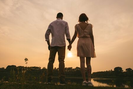 Couple standing hand by hand on a river shore, watching a beautiful sunset taking place. Low angle. Man has a prosthetic bionic arm.の写真素材