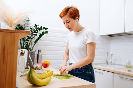 Emotionless focused middle aged woman with short ginger hair slicing banana for a muesli breakfast in the kitchen. She's wearing casual clothes: white T-shirt and jeans.の写真素材