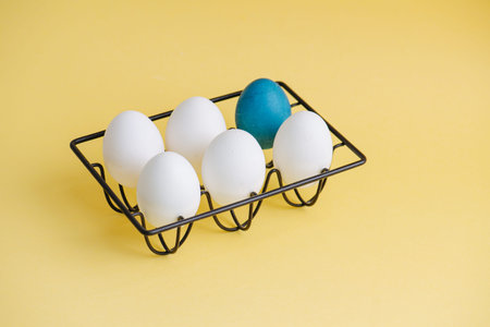 White eggs and one blue in a metal stand on a pastel yellow background