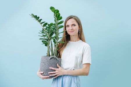 Happy pleased woman standing with a plant in a wicker pot, she's holding in her hands, over blue background. She has an extra long beautiful hair and casual clothes.の写真素材