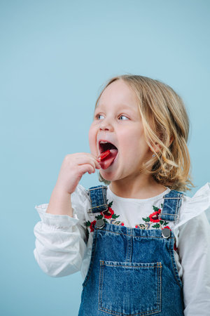 Funny little blonde girl in jeans overalls eating lollipop candy with her mouth wide-open. Looking...