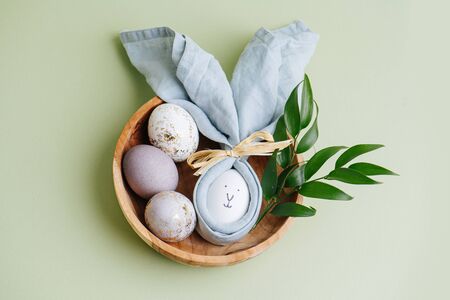 Easter egg in a napkin with a rabbit face painted on it along with gilded eggs in a wooden bowl and plant branch. Over pale green surface. Top view.の写真素材
