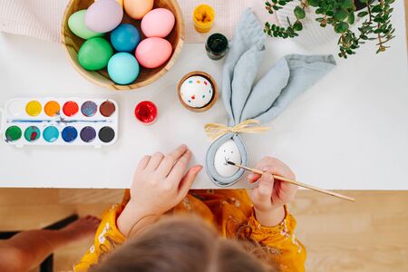 Little brunette girl in orange shirt sitting behind a table, painting face on easter egg in a napkin, using brush. Top view.の写真素材