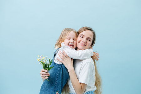 Little girl receives a hug from her mother for giving bouquet of wild camomiles. Both look happy. Over blue background.の写真素材