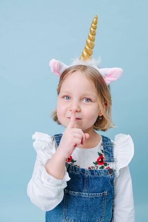 Cute little blonde girl in unicorn head band and jeans overalls making silence gesture over blue background. Pressing index finger against her lips.の写真素材