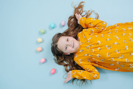 Happy little brunette girl in orange shirt lies on a blue-surfaced floor with a rainbow of easter...