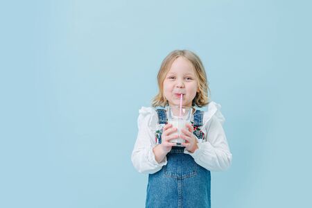 Cute cheerful little blonde girl in overalls and fluffy white shirt drinking milkshake with a straw, holding glass with both of her tiny hands over blue backgroundの写真素材