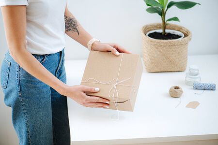 The girl wrapped a gift in a beautiful white interior near the table and holds it in her hands in a stylish craft. Close-upの写真素材