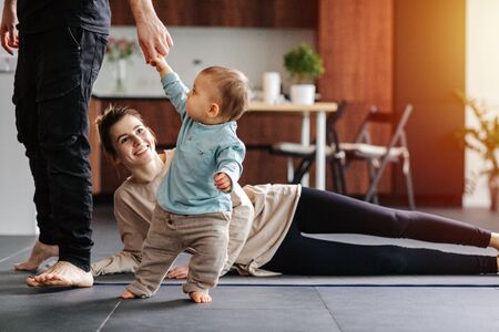 Mother doing side leg raises while father walks around, carrying their infant baby in his hands. She's amused, her eyes closed. Family quarantine, domestic life in self-isolation.の写真素材