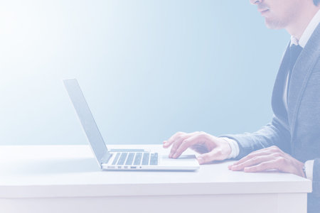A young business man in a jacket and tie sits at a white desk and works behind a laptop, touches a...