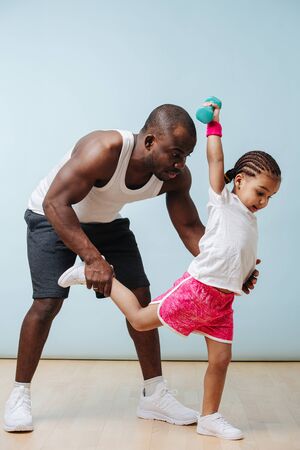 Father working out with his daughter. He's holding her by the leg while she tries to maintain balance with dumbbell in a hand, outstreched upwards.の写真素材