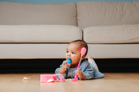 Little infant baby playing on the floor at home in front of the couch, stuffing doll head in her mouth. Lying on her stomach, head is lifted.の写真素材