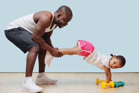 Father and daughter are working out during lockdown. He holds her by the legs, she's holding plank.の写真素材