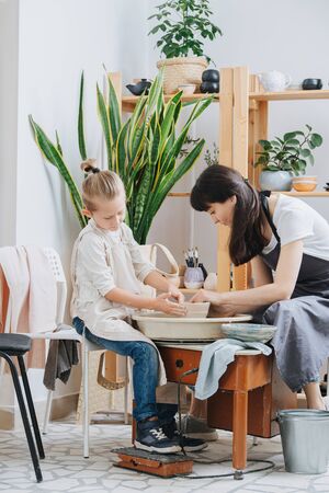 MIddle age brunette woman teaching little blond boy how to shape clay tableware on a pottery wheel in a private workshop. Chance to spend time on learning new skills during isolation.の写真素材