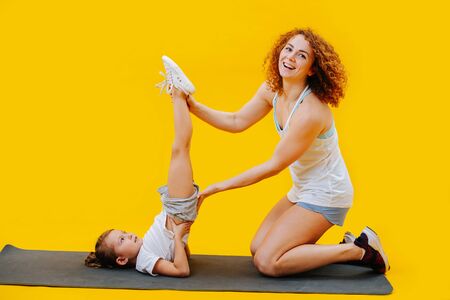 Mother and daughter doing candlestick stand with leg holding assistance on a yoga mat, synchronous over yellow backgroundの写真素材
