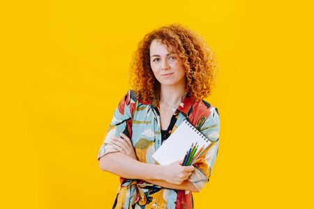 Happy beautiful young woman in a colorful dress with red curly hair with notepad and crayons in hands over yellow. Smiling at the camera.の写真素材