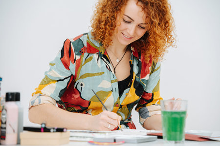 Smiling artist woman painting behind work desk in her notepad with a brush