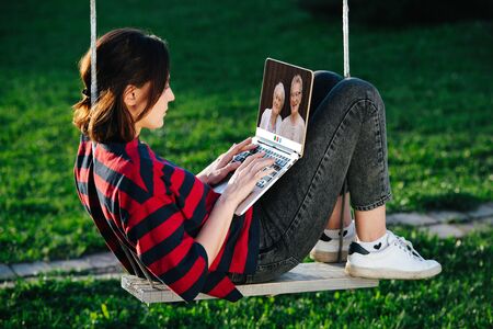 Girl sitting on a swing with a laptop in her hands talking by video link with her grandparentsの写真素材