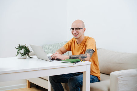 Happy bold freelancer in glasses working on his laptop behind the table at home during covid-19...