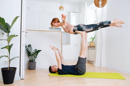 Adult siblings doing gymnastics together during isolation at home. Guy lying on his back on a mat, holding girl in the air with straight legsの写真素材
