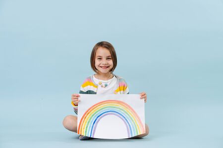 Cheerful little girl showing a rainbow she just painted. Sitting on the floor. Over blue background.の写真素材