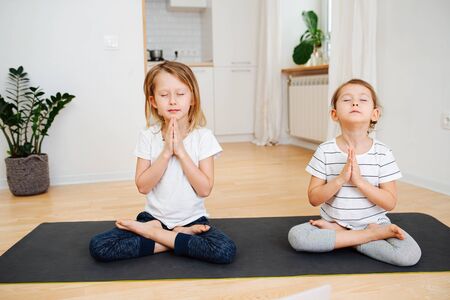 Two little siblings doing yoga on a mat at home, sitting in full lotus pose with joined hands and closed eyes.の写真素材