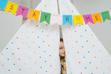 Playful little girl peeking from a hut, made with sticks and bedsheets. Stay at home flag garland hanging across overhead.の写真素材