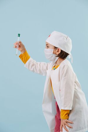 Little girl playing a doctor over blue background. Pandemic inspired. Child dreams to be a doctor to save peoples lives from coronavirus. Holding test-tube with colored liquid.の写真素材