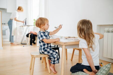 Children eating behind a small table, their mom ironing clothes in the blurred backgroundの写真素材
