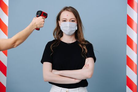 Portrait of a young girl in a protective mask, stands at the epidemic control point, staff measures her temperatureの写真素材