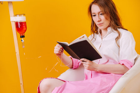 Young sick woman reading while waiting for her infusion drip bottle to go emptyの写真素材