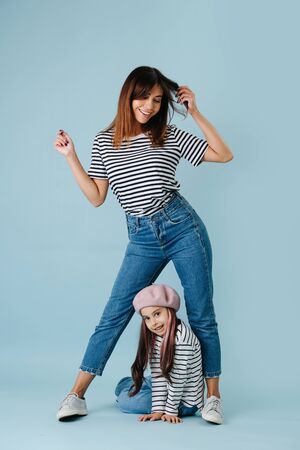 Two fashion lovers, mom and daughter wearing same clothes over blue background. They have jeans and black and white striped shirt on them. Mom in a flattering pose, looking down at her daughter.の写真素材