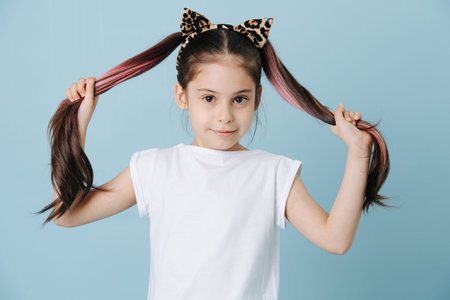 Portrait of a cute little girl with cat ears headband holding her ponytails in hands, posing for a...