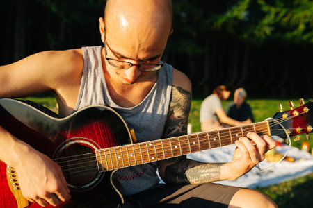 Young bold man sitting with guitar, looking down at it, learning to play during spring picnic with...