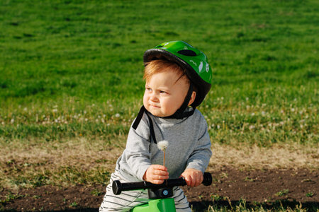 Grumpy little kid on a three-wheel bike in a countryside. He's holding a dandelion. Wearing protective helmet. Over field of grass.の写真素材